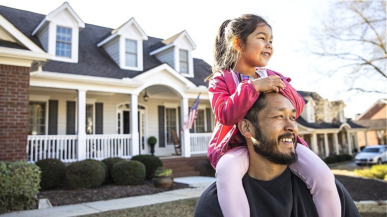 Father carrying daughter on shoulders smiling in front of house