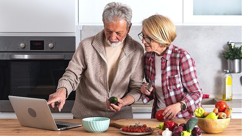 Older couple reviewing laptop while cooking in kitchen