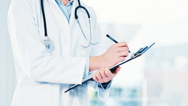 A person wearing a stethoscope and a white lab coat holding a clipboard and a pen.