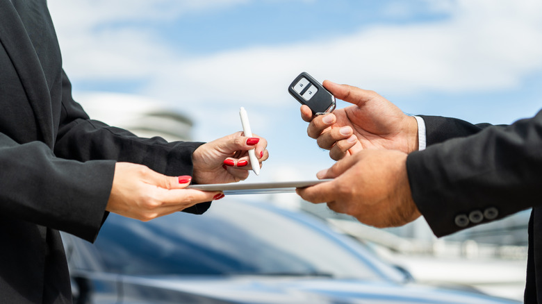 Person signing paperwork while another hands them car key.