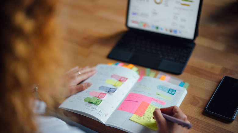 A curly-haired person fills out a paper planner while looking at an iPad pro and a cell phone.