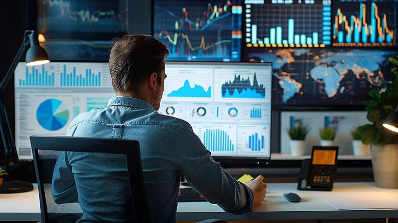 A man seated in front of screens displaying graphs and charts