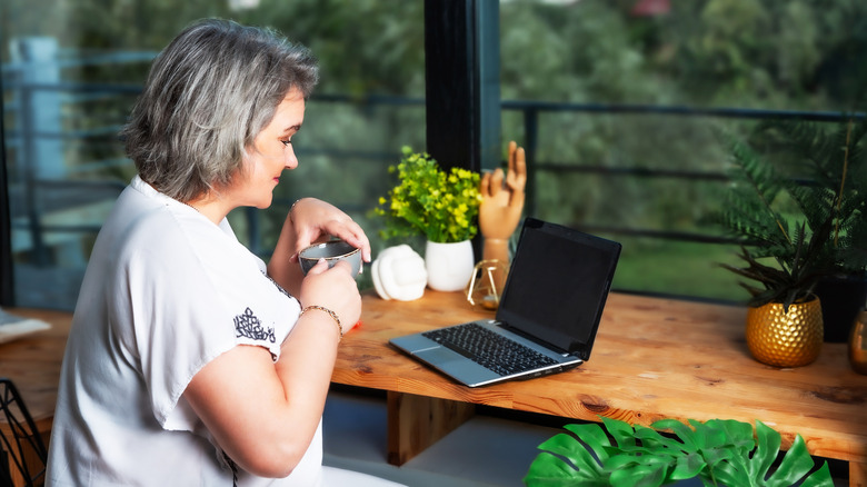 Woman works on laptop sitting in cozy room near window with cup