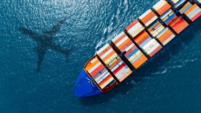 A plane shadow against the ocean, featuring a large container ship full of colorful shipping containers on it.