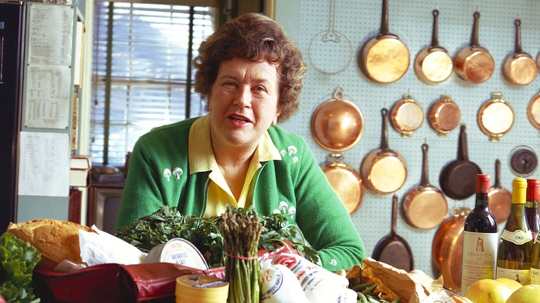 Julia Child in green sweater posing behind counter that's filled with food