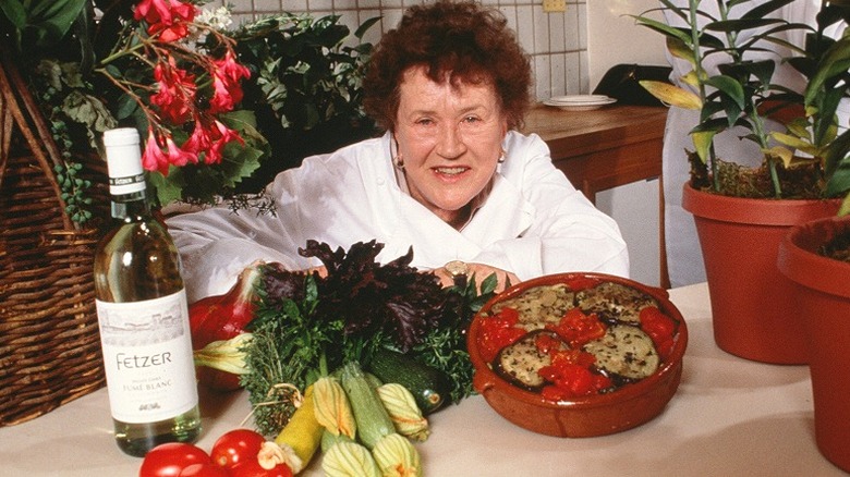 Julia Child smiling behind a display of food, surrounded by plants