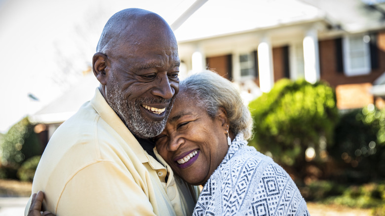 A smiling elderly couple hugging with a house in the background