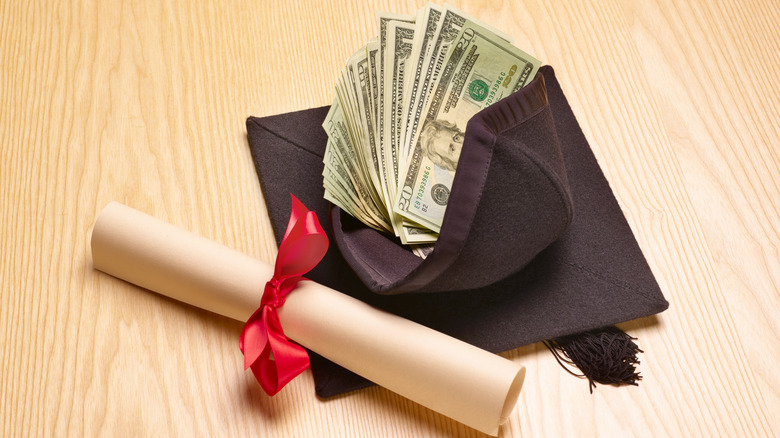 A rolled-up diploma next to a graduation cap filled with money.