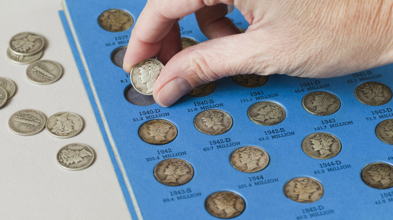 A hand placing an old coin into a page of a coin collecting book with other coins already placed in slots on the page.