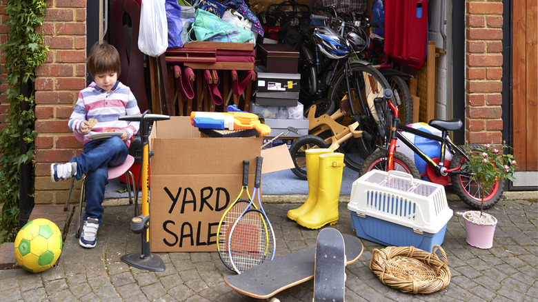 A small child siting among a number of things for sale at a yard sale.