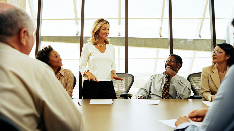 A female executive standing in a boardroom with smiling employees seated at a table