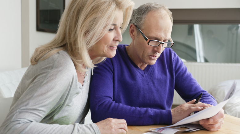 A man with grey hair, glasses, and wearing a blue sweater sitting next to a woman with blond hair wearing a grey sweater are seated at a table looking at a piece of paper together.