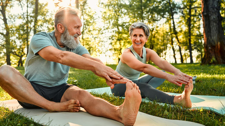 An adult man and an adult woman sitting outdoors on yoga mats stretching.