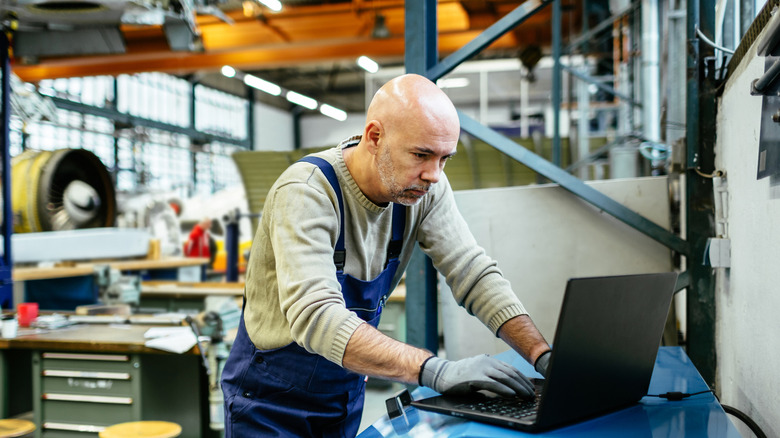 A Gen X man in an industrial warehouse typing on a laptop