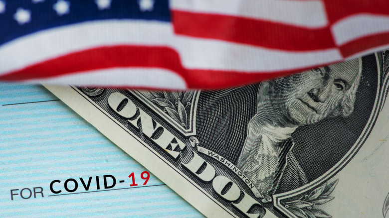 An American flag waves over a single dollar bill and a sliver of a stimulus check for COVID-19.