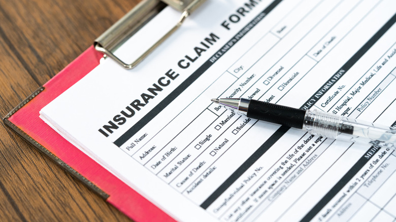 A red clipboard holding a blank insurance claim form and ballpoint pen on a wooden table.