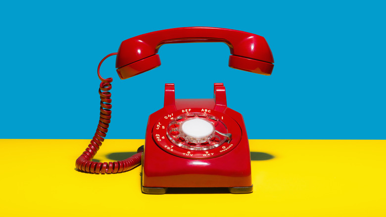 An old-fashioned red rotary phone against a bright blue and yellow colorblocked background.