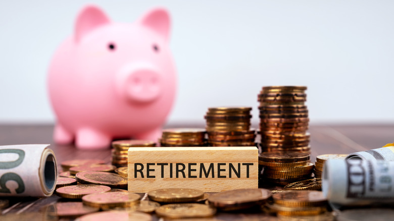 The word 'Retirement' on a small wooden sign surrounded by piles of coins, rolls of bills, and a pink piggy bank in the background.