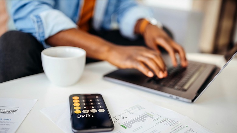 Person typing on laptop at home with coffee, calculator, and paperwork on table