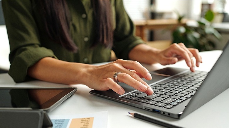 Close-up of a person typing on a laptop with a tablet, pencil, and paperwork in view