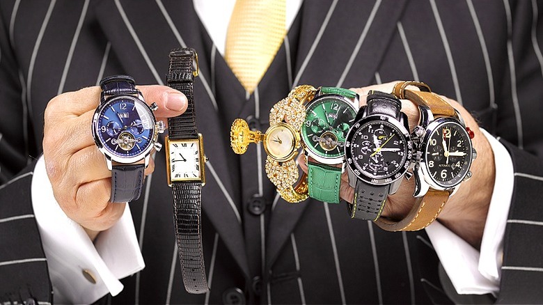 A person wearing a black suit with gray pinstripes, a while shirt, and yellow tie holds a variety of watches in various colors and styles in both hands.