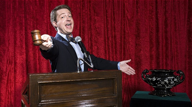 A man standing at a podium with a microphone holding a wooden gavel in one hand and pointing at a black and white vase with his other hand.