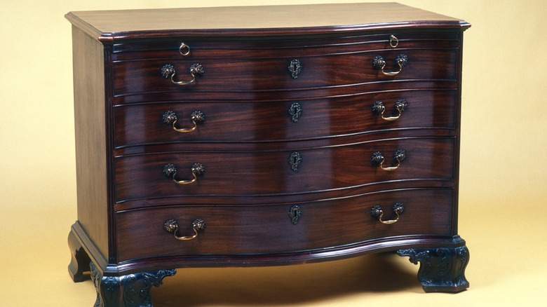 A very recognizable type of dark brown Chippendale chest of drawers against a yellow background.