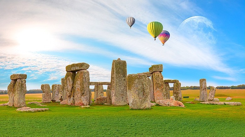 A trio of colorful hot air balloons float over Stonehenge, in front of a daytime blue sky with the moon visible