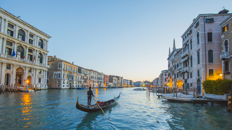 A gondolier rows down an otherwise empty canal in Venice at twilight, among the glittering piers and palazzos.