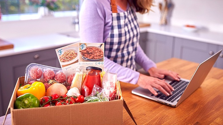 Open meal kit waits as woman researches something on her laptop