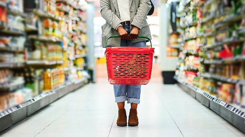 Woman stands in the middle of a grocery aisle holding shopping basket