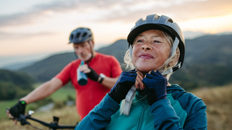 Senior couple gets ready for a mountainous bike ride, sipping water and strapping on a helmet.