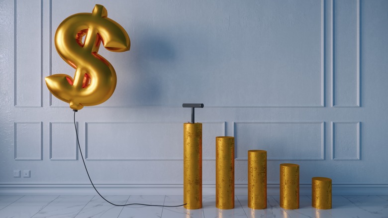 A gold inflated money sign balloon hovers over a stack of gold coins to represent inflation.