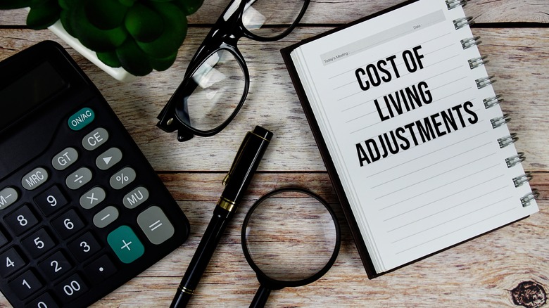 A notebook with "Cost of Living Adjustments" stamped on its open page, next to a pair of glasses, pen, calculator, and magnifying glass on a wooden picnic table.