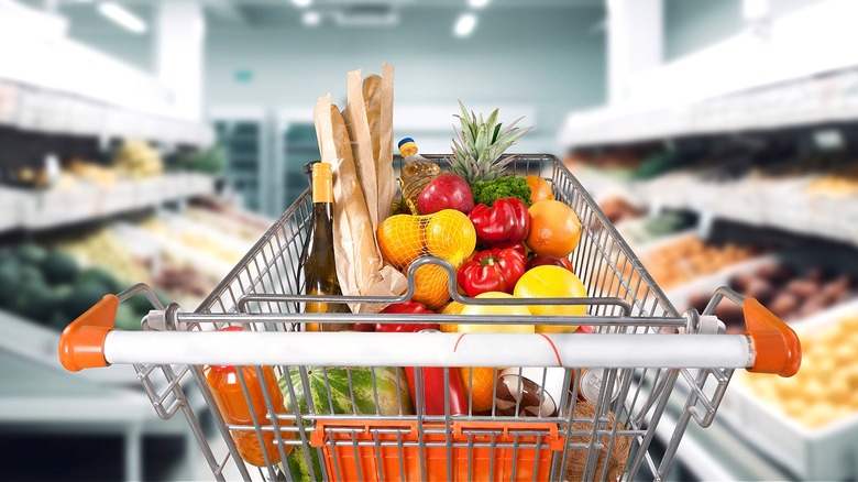 A shopper's POV of a produce-laden shopping cart in a brightly lit grocery store