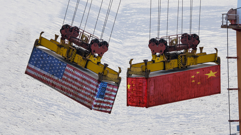 A shipping container emblazoned with the American flag nearly bonks a shipping container emblazoned with the Chinese flag as the two containers dangle over port.