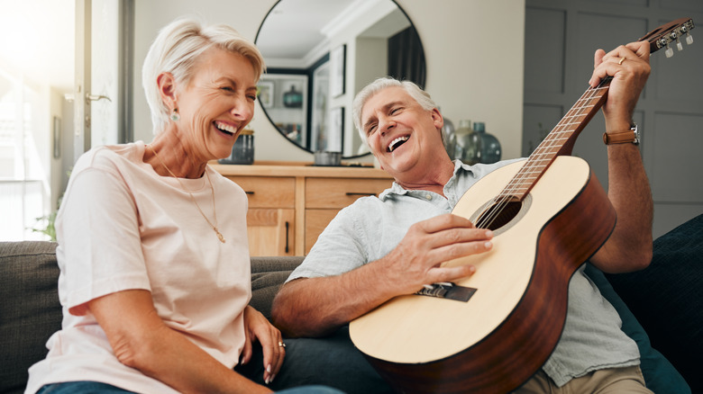 An elderly man and woman sitting on a sofa laughing while the man plays guitar