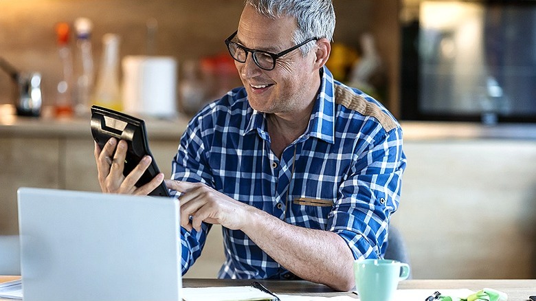 Man smiling while using calculator at table with laptop, coffee cup, and paperwork