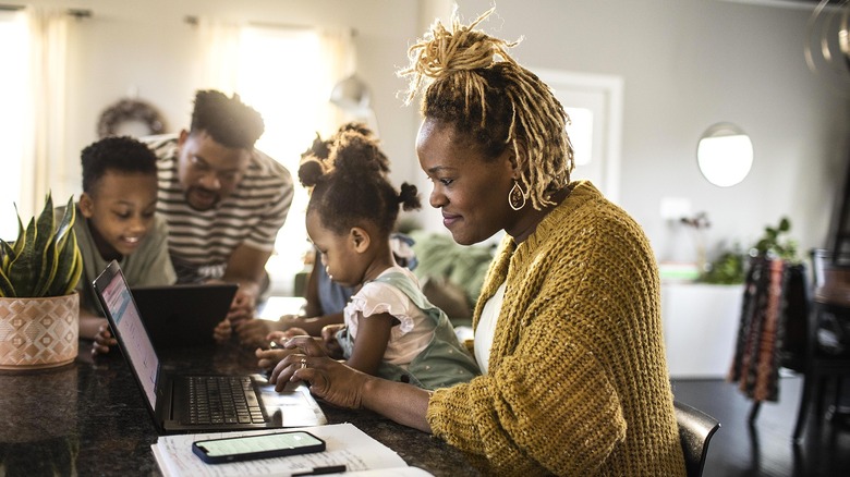 A father and kids use a tablet while a mother holds her baby and searches on a laptop