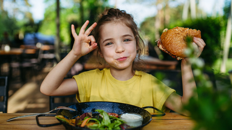 A young girl holding a burger and smiling on a restaurant patio