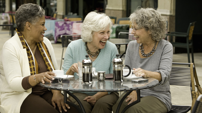 Three laughing senior women seated on a patio with coffee