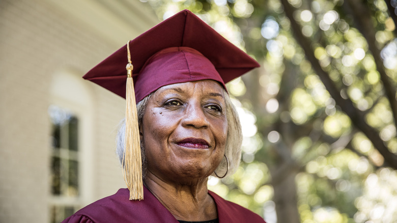 A smiling senior woman wearing a graduation gown and cap outside.