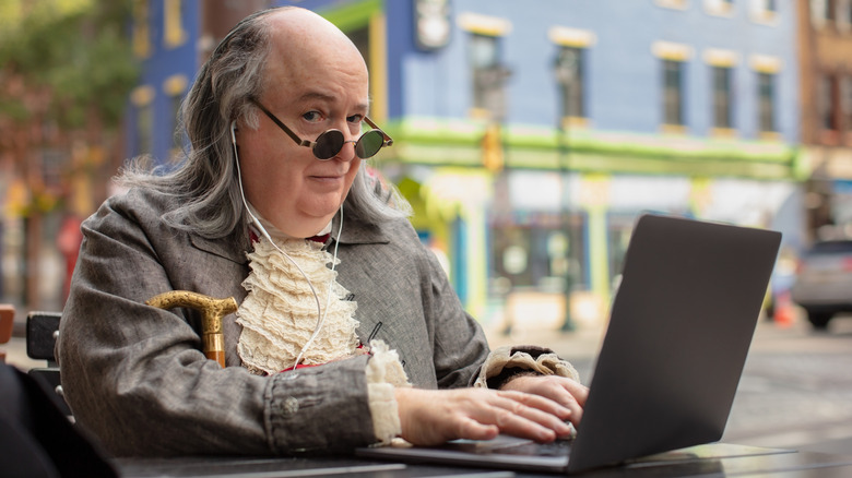 A senior male in period costume seated outside typing on a laptop