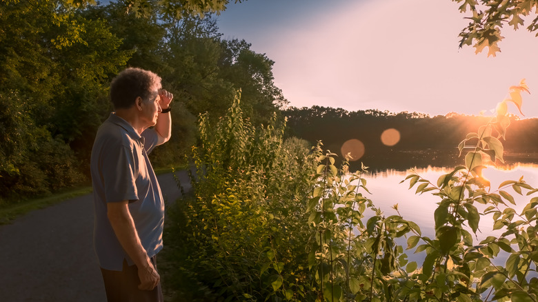 An elderly man standing on the side of road watching a sunset over a lake