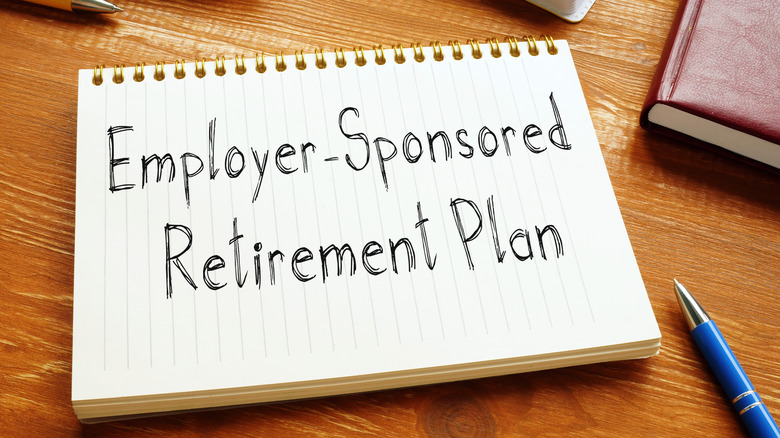 A notebook open to a page with the words 'Employer-Sponsored Retirement Plan' written on it sitting on a wooden desktop surrounded by pens and books.