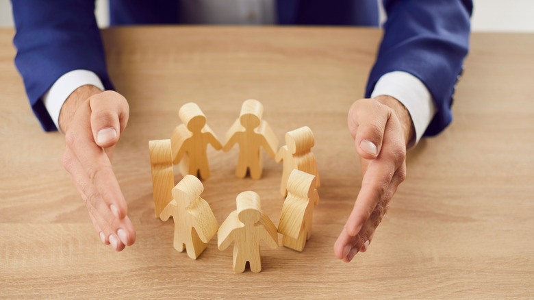 A group of wooden human-like figurines arranged in a circle with a pair of hands surrounding them on either side.