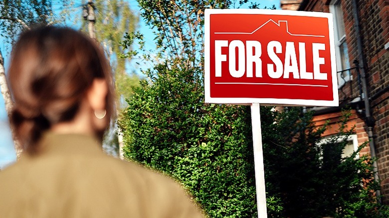 Woman looking up at a 'For Sale' sign in front of brick home