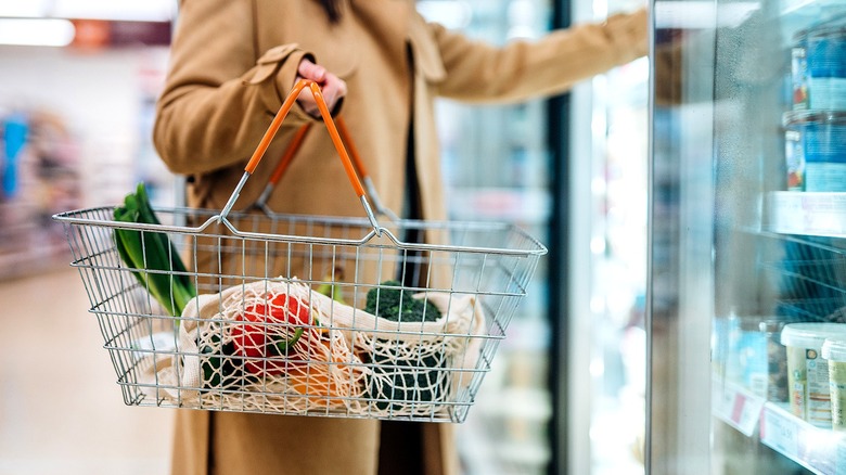 Person looks in grocery store freezer while holding metal shopping basket containing produce and a bag