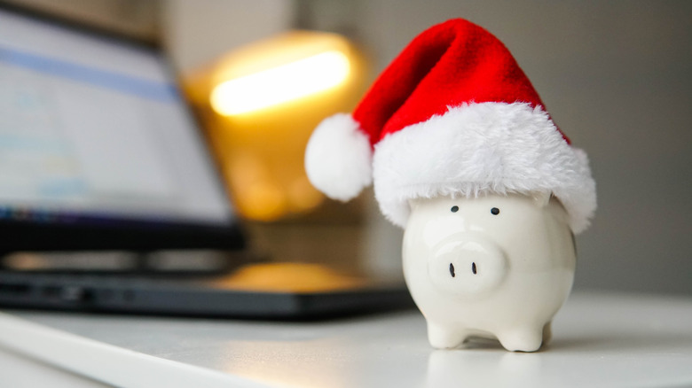 A small white piggy bank wearing a red Santa hat with a blurred image of a laptop in the background.