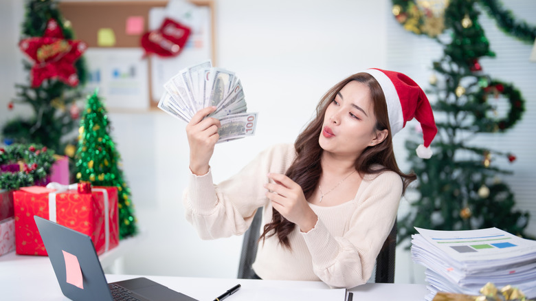 A young woman wearing a Santa hat sitting at her desk holding a handful of cash with Christmas decorations in the background.